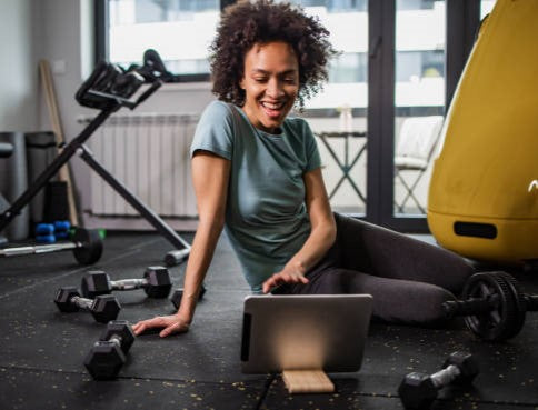 en kvinna sittandes på golvet intill träningsredskap med en dator framför sig under samtal med sin personliga tränare